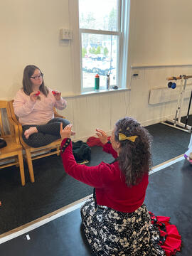 Miss Coco seated on floor teaching a seated female high school student with Downs Syndrome how to play castanets