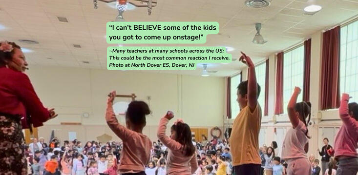 Four students with arms up onstage with Miss Coco in front of large grade school audience.